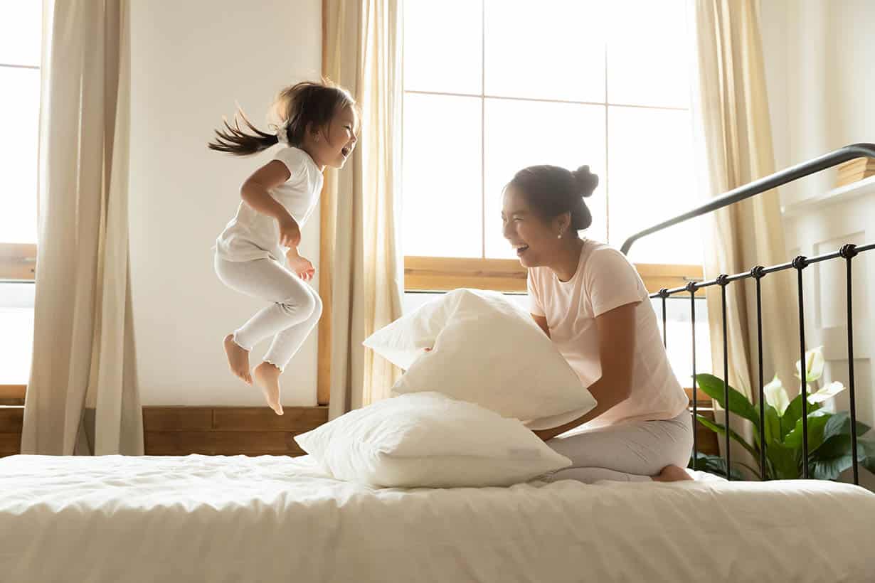 Carefree small daughter jump on bed while vietnamese mother laughing